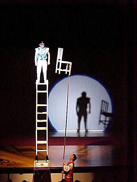 A performer stands on a tower of chairs, and is offered another.